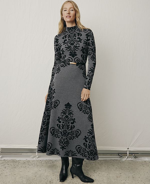 A woman wearing a long-sleeved, grey and black patterned dress with black boots stands against a plain white backdrop.