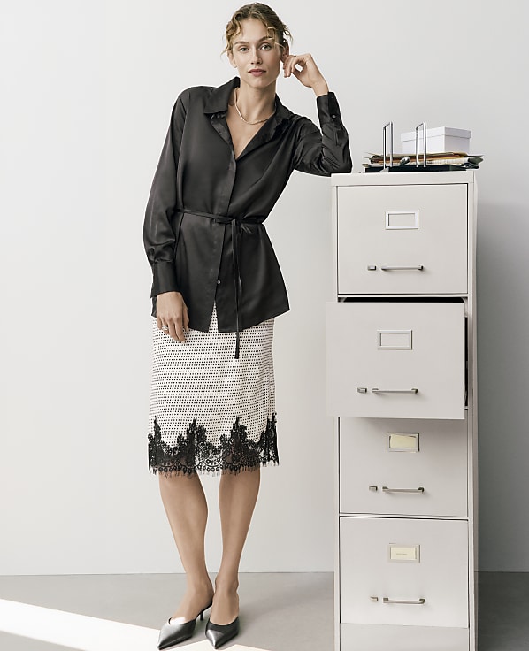 A woman wearing a black satin shirt and a patterned lace skirt leans against a tall white filing cabinet in a minimalist white room.