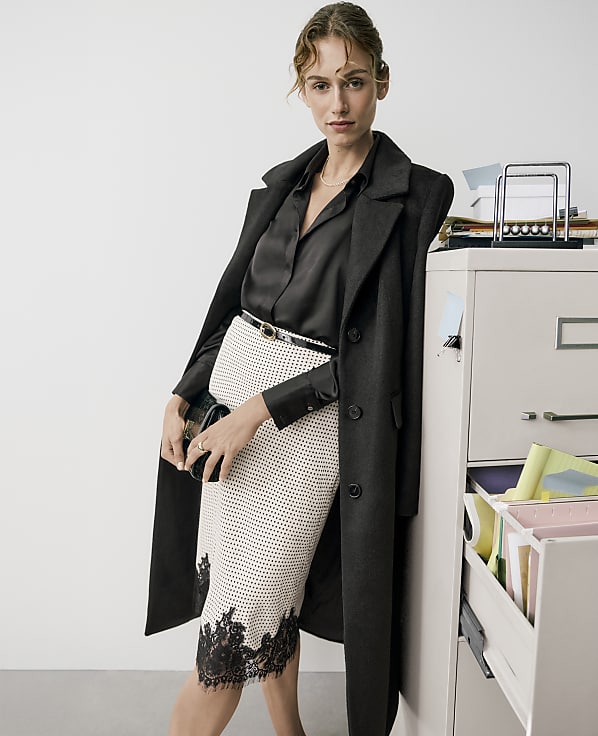 A woman wearing a black coat and a patterned skirt leans against a white filing cabinet in a minimalist white room.
