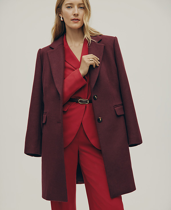 A blonde woman wearing a red suit and a burgundy coat stands against a plain light gray background.