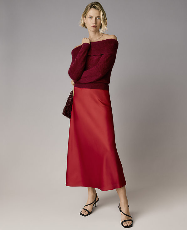 A blonde woman wearing a burgundy off-the-shoulder sweater and a red satin skirt stands against a plain grey background.