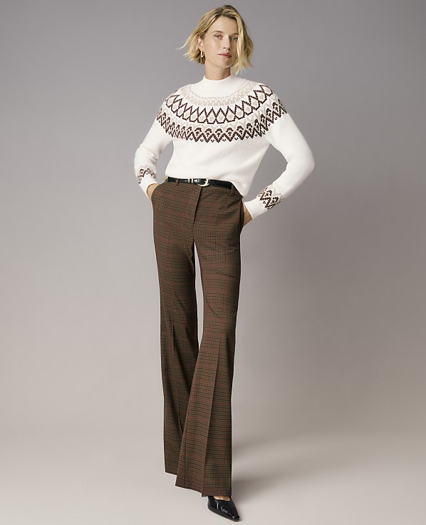 A woman wearing a white patterned sweater and brown plaid trousers stands against a plain grey background.