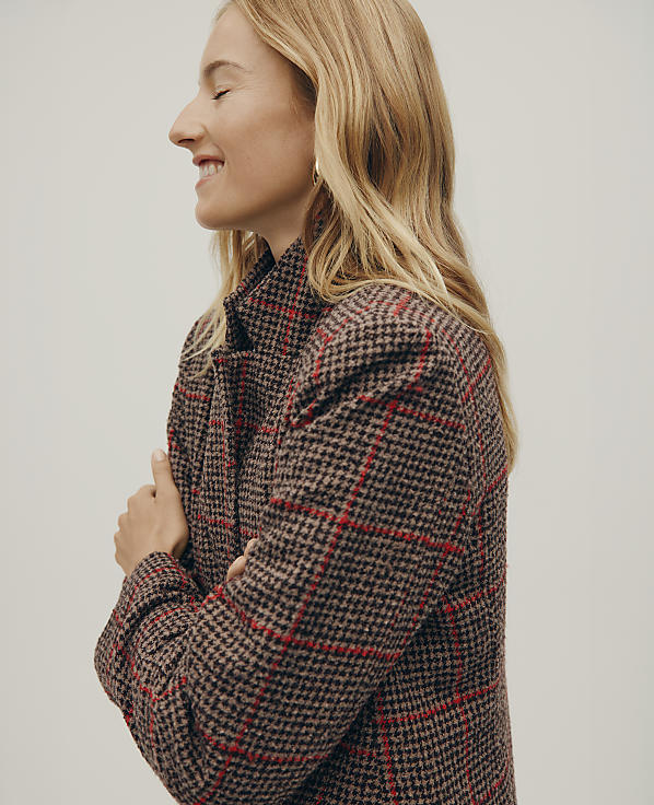 A smiling blonde woman in a brown and red plaid coat stands against a plain, light-colored background.