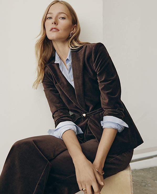 A blonde woman wearing a brown velvet suit and light blue shirt sits against a plain white wall.