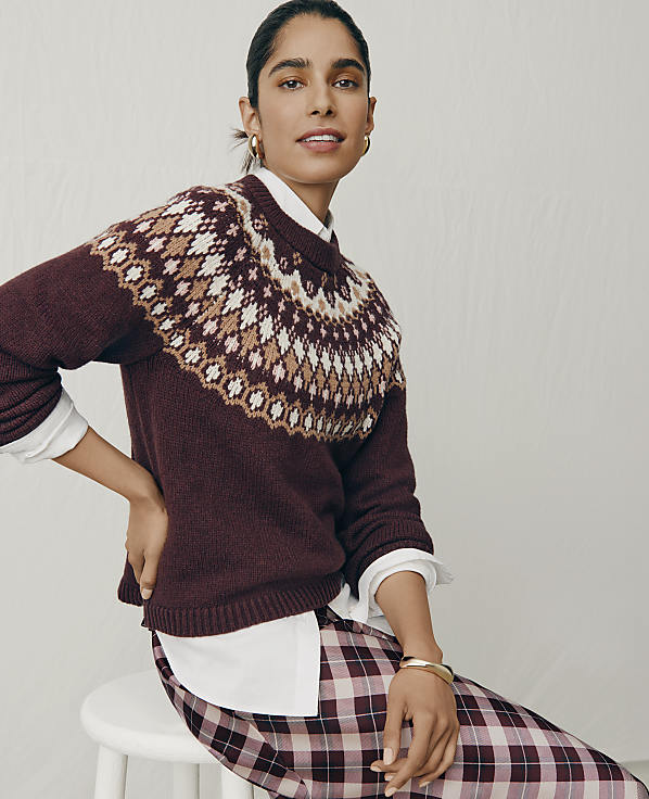 A woman wearing a maroon patterned sweater and plaid pants sits on a white stool against a plain light gray background.