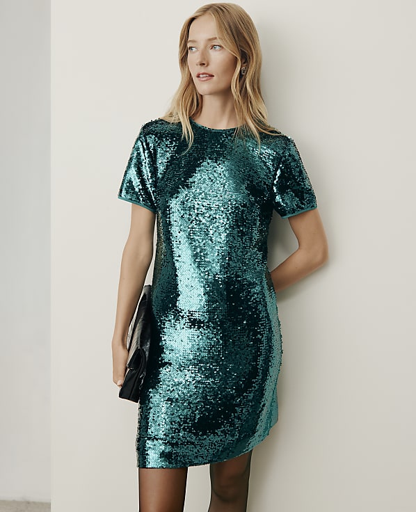 A woman wearing a shimmering teal sequin dress and holding a black clutch stands against a plain, light-colored wall.