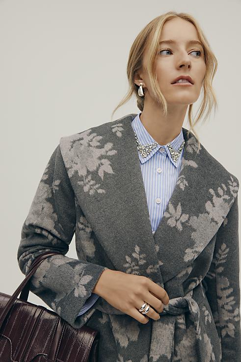 A blonde woman wearing a grey floral coat and a blue striped shirt stands against a plain, light-colored background.