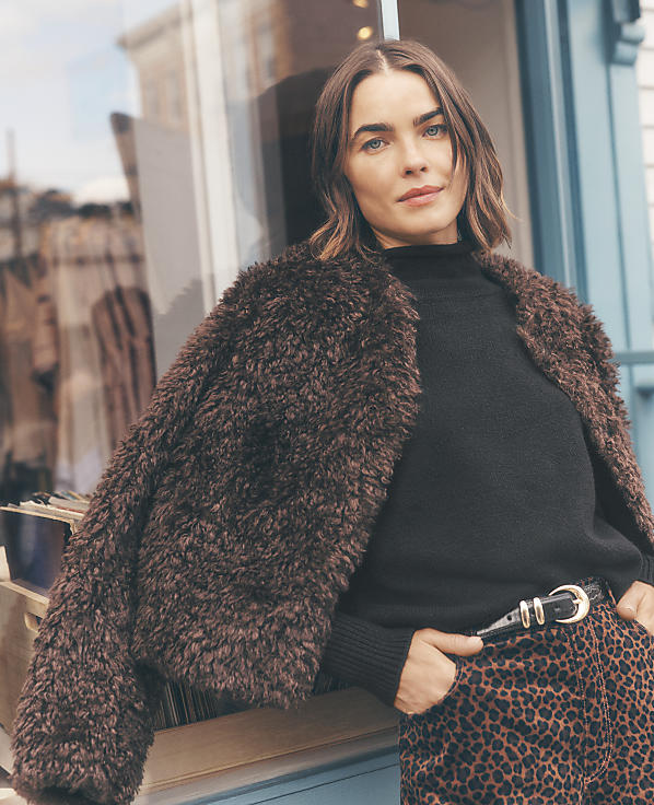 A woman wearing a brown faux-fur coat, black sweater, and leopard-print pants leans against a storefront window.