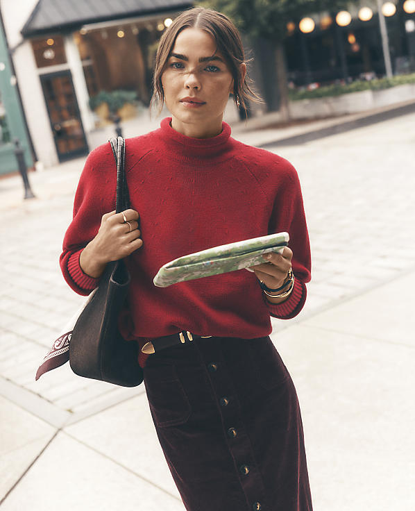A woman wearing a red sweater and burgundy skirt stands on a city sidewalk with blurred storefronts in the background.
