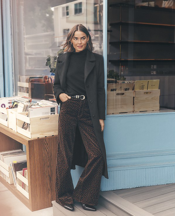 A woman wearing a black coat and leopard-print trousers stands in front of a blue storefront window filled with crates of vinyl records.