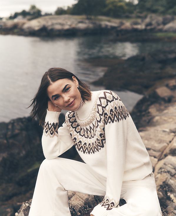 A woman in a white patterned sweater and white pants sits on a rocky shore with a calm body of water and distant greenery in the background.