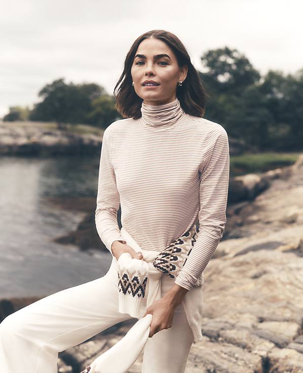 A woman wearing a light-colored turtleneck and white pants stands on a rocky shoreline with a calm body of water and green trees in the background.