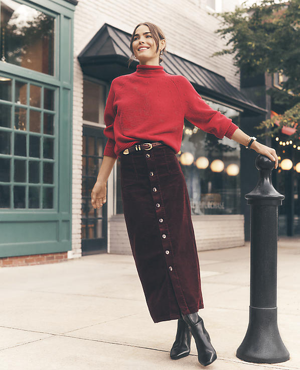 A smiling woman in a red sweater and long burgundy skirt leans against a black bollard on a city sidewalk in front of storefronts.