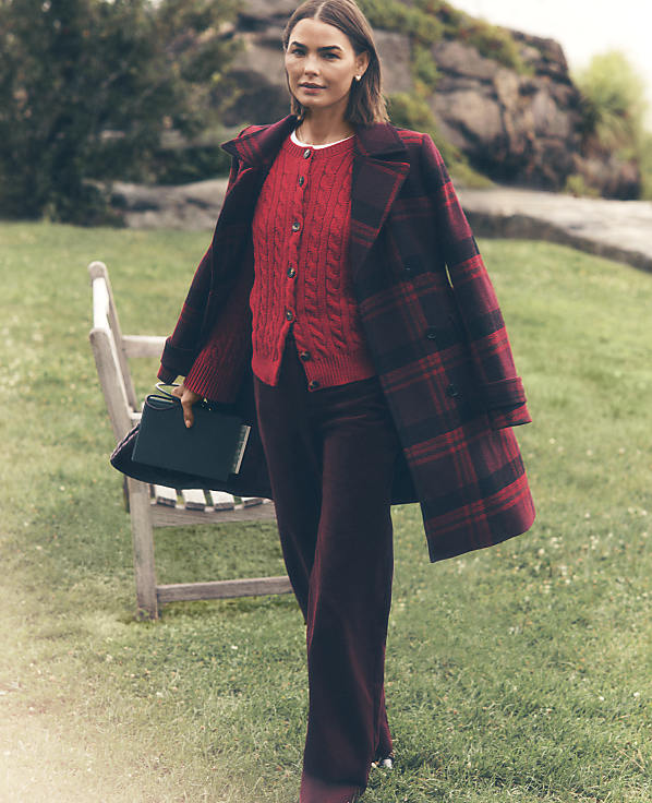 A woman wearing a red cardigan and a red and black plaid coat stands on a grassy lawn with a wooden bench and rocky terrain in the background.