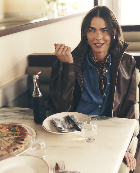 A woman in a leather jacket and leopard print scarf sits at a marble table with pizza and drinks in a cozy restaurant booth.