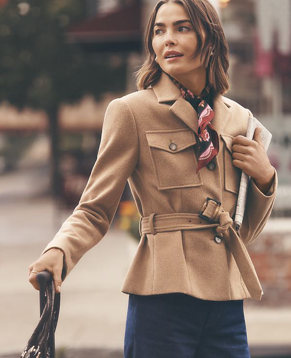 A woman wearing a belted tan jacket and a patterned scarf stands in the foreground against a blurred city street background.
