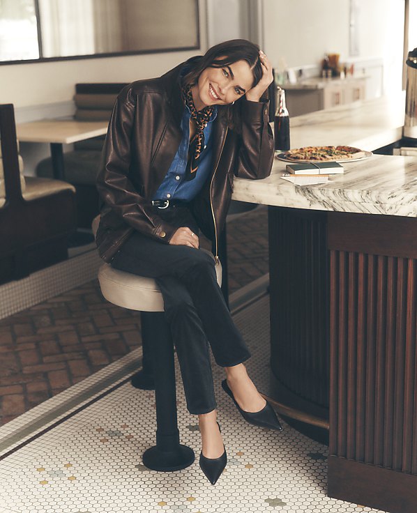 A woman in a brown leather jacket and blue shirt sits on a stool at a marble counter, with a vintage-style diner interior in the background.