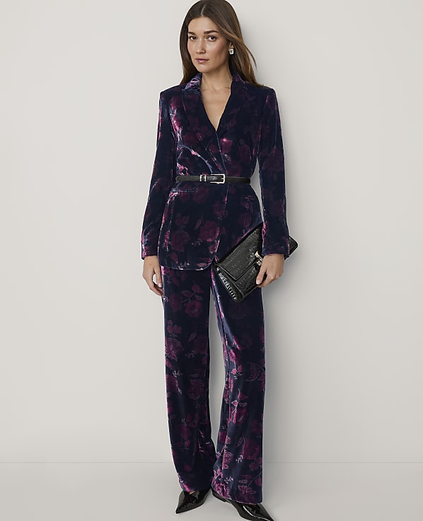 A woman wearing a dark purple velvet floral suit and holding a black clutch stands against a plain white background.