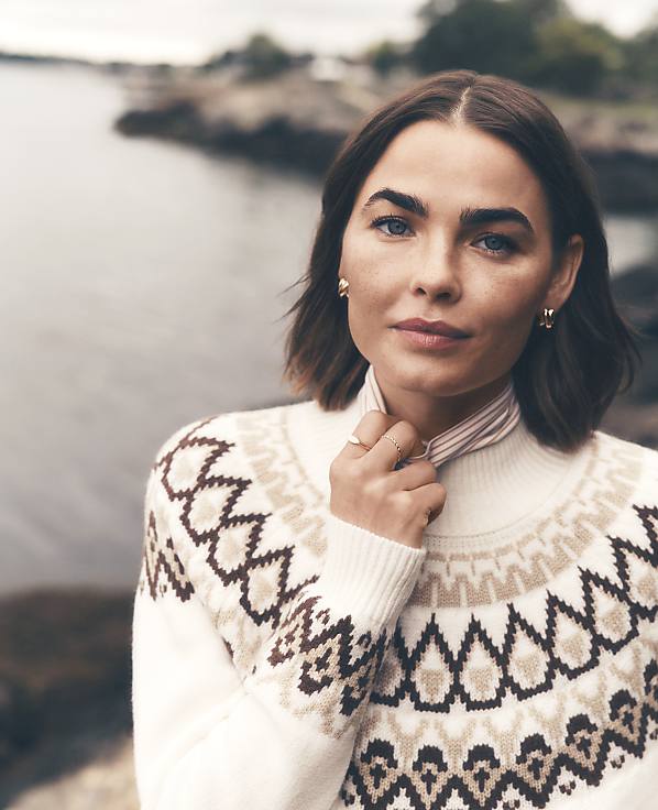 A woman wearing a patterned white sweater stands in the foreground against a blurred background of a rocky coastline and water.