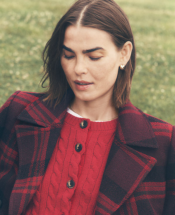 A woman wearing a red cable-knit sweater and a red and black plaid coat stands against a blurred green grassy background.
