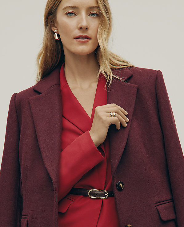A blonde woman wearing a red dress and a burgundy coat stands against a plain, light-colored background.
