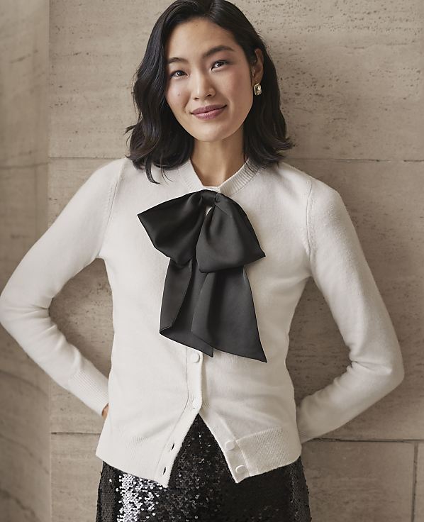 A woman wearing a white cardigan with a large black bow and a sequined skirt stands against a neutral stone wall.