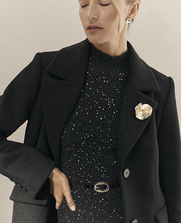 A woman wearing a black coat and a shimmering black sequined dress stands against a plain, neutral-toned background.