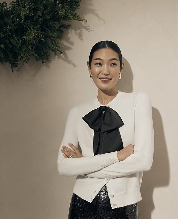 A woman in a white cardigan with a black bow stands with crossed arms against a neutral wall with greenery in the upper left corner.