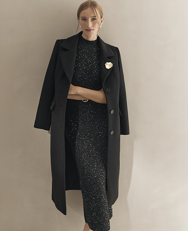 A woman wearing a shimmering black dress and a long black coat stands against a plain, neutral-toned wall.