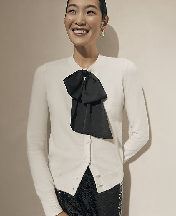 A smiling woman wearing a white cardigan with a large black bow and sequined bottoms stands against a plain, neutral-toned wall.