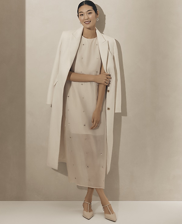 A smiling woman wearing a beige dress, a white coat, and pointed heels stands against a plain, neutral-toned wall.