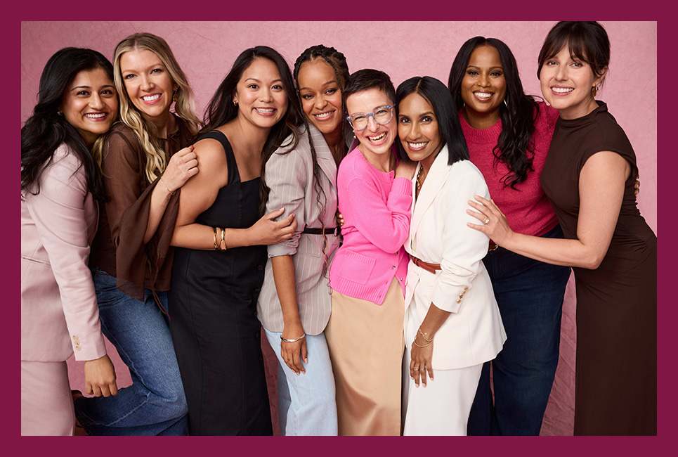 A diverse group of eight women stand closely together, smiling and posing against a plain pink background.