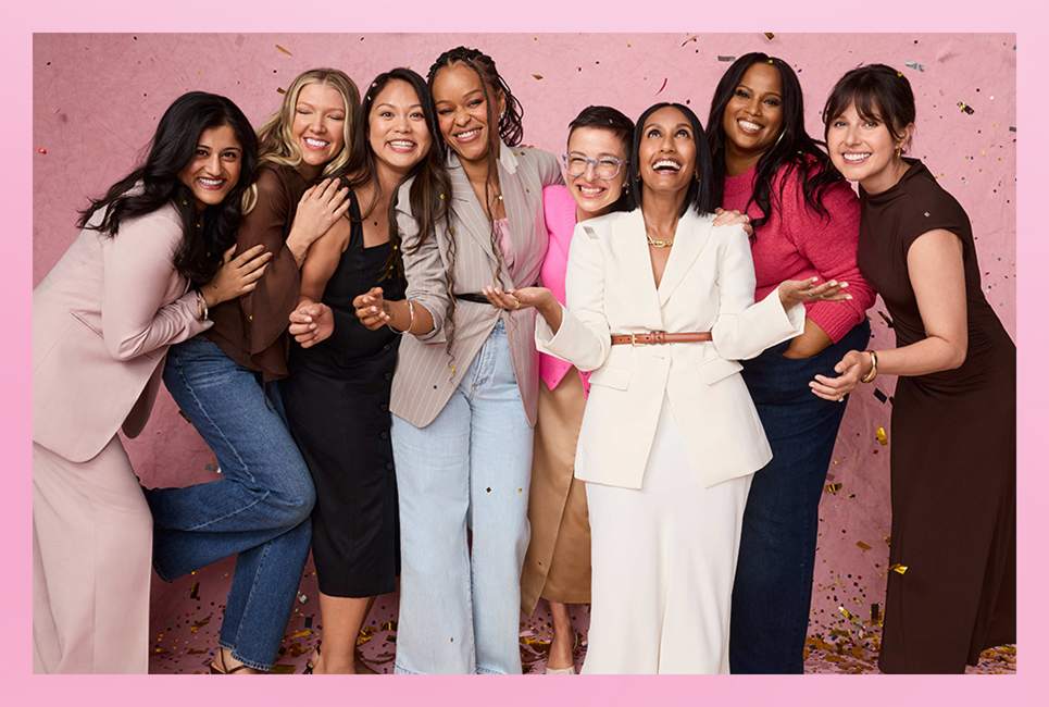 A diverse group of eight women stand closely together, laughing and smiling against a pink background with confetti.